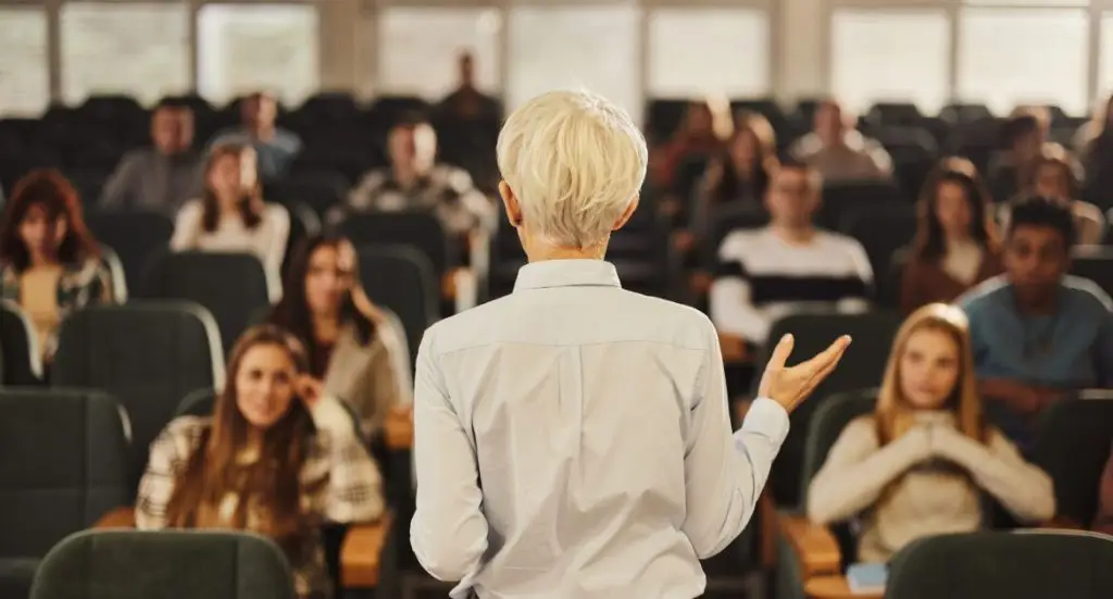 "A lecturer, seen from behind, addressing a large, seated audience of students in a lecture hall or classroom."